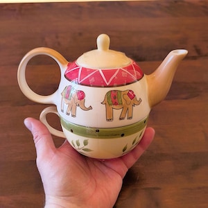 May include: A cream-colored teapot and teacup set with elephant designs. The teapot has a red and white geometric pattern on the lid and a green band with leaves. The elephants are tan with colorful accents.