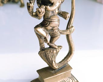 May include: A gold-colored metal statue of the Hindu deity Ganesha. The statue depicts Ganesha standing on a lotus flower and holding a snake in his trunk. The statue is on a square base with intricate carvings.