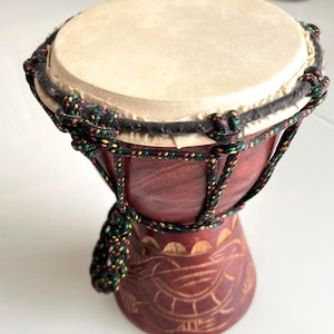 May include: A small, handcrafted djembe drum with a light tan drumhead and a dark reddish-brown wooden body. The drum is adorned with a decorative rope in black, green, and yellow. The base of the drum features carved patterns.