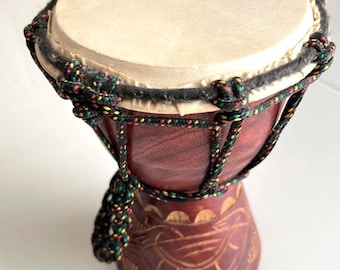 May include: A small djembe drum, handcrafted with a light tan drumhead and a dark reddish-brown wooden body. The drum is decorated with a rope in black, green, and yellow. The base of the drum has carved patterns.