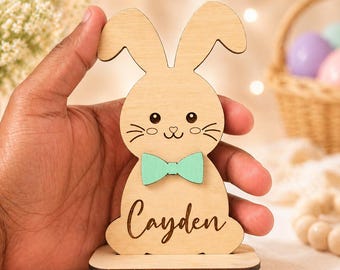 May include: Wooden Easter bunny decoration with a mint green bow tie and the name "Cayden" engraved on the front. The bunny has a cute face with whiskers and is standing on a small wooden base. A basket of Easter eggs is in the background.