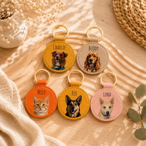 May include: A collection of personalized pet keychains. Each keychain features a circular design with a gold-colored ring and a leather-like backing. The keychains display images of a cat and dogs, with the pet's name printed above the image.