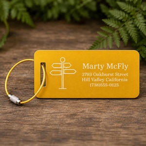 May include: Mustard yellow luggage tag with a directional sign graphic and the name "Marty McFly". The tag includes an address and phone number. A gold-coloured wire loop with a silver clasp is attached.