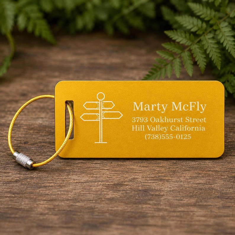 May include: Mustard yellow luggage tag with a directional sign graphic and the name "Marty McFly". The tag includes an address and phone number. A gold-coloured wire loop with a silver clasp is attached.