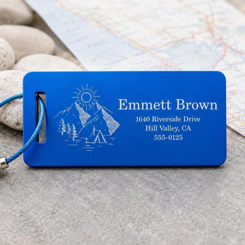 May include: A blue luggage tag featuring a mountain and sun design, the name "Emmett Brown", and address details. It has a metal loop and a blue wire cable. The background shows a map and small stones.
