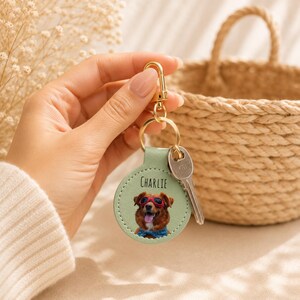 May include: A round, light green leather keychain with a gold clasp and key. The keychain features a dog wearing sunglasses and a blue scarf, with the name "Charlie" printed above the dog's image.
