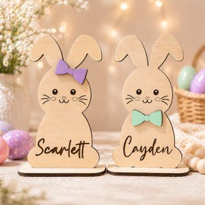 Op de afbeelding: Twee houten paashaasdecoraties. E&eacute;n konijn heeft een paarse strik en de naam "Scarlett" erop geschreven. Het andere konijn heeft een groene strik en de naam "Cayden" erop geschreven. De konijnen staan op een houten voet.