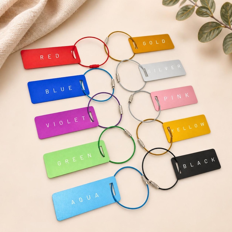 May include: A selection of brightly coloured luggage tags with metal loops. Each tag is labelled with a colour name: red, blue, violet, green, aqua, gold, silver, pink, yellow, and black. The tags are rectangular and shiny.