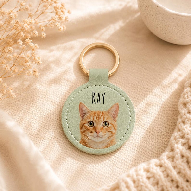 May include: A round, light green keychain with a gold-colored ring. The keychain features a photo of an orange tabby cat with the name "RAY" printed above the cat's face. The keychain is made of a leather-like material.