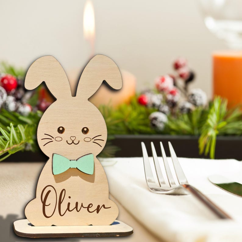 Op de afbeelding: Houten paashaas plaatskaart met een mintgroene vlinderdas en de naam "Oliver" gegraveerd. De haas is lichtbruin en heeft een schattig gezicht. Voor tafeldecoratie.
