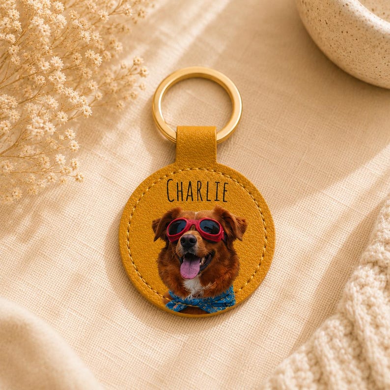 May include: A round, mustard-yellow leather keychain with a gold ring. The keychain features a dog wearing sunglasses and a blue bandana. The name "Charlie" is printed above the dog's image. The keychain is a personalized pet accessory.