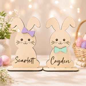 Op de afbeelding: Twee houten paashaasdecoraties. E&eacute;n konijn heeft een paarse strik en de naam "Scarlett" op de basis. Het andere konijn heeft een zeegroene strik en de naam "Cayden". Beide hebben schattige snorharen en ogen.