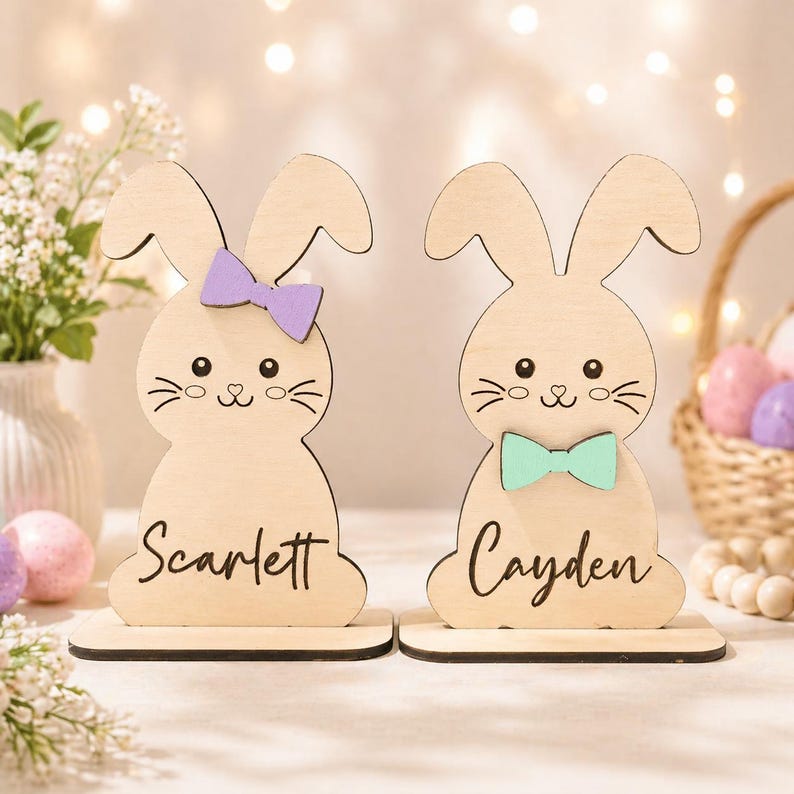Op de afbeelding: Twee houten paashaasdecoraties. E&eacute;n konijn heeft een paarse strik en de naam "Scarlett" op de basis. Het andere konijn heeft een zeegroene strik en de naam "Cayden". Beide hebben schattige snorharen en ogen.
