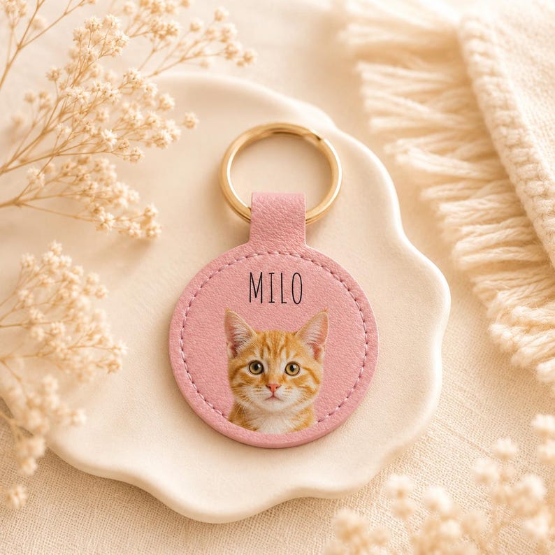 May include: A pink leather keychain with a gold ring. The keychain features a circular design with a photo of an orange tabby cat and the name "MILO" printed in black.