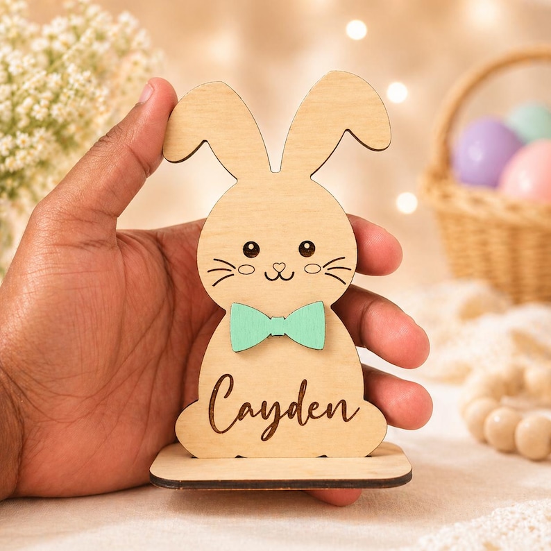 Op de afbeelding: Houten paashaasdecoratie met een mintgroene strik en de naam "Cayden" gegraveerd. De haas heeft een schattig gezicht met snorharen en staat op een kleine houten basis. Een mand met paaseieren is op de achtergrond.