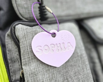 May include: A purple heart-shaped tag with the name "Sophia" written in a wavy font. The tag is attached to a purple cord and is hanging from a grey backpack.