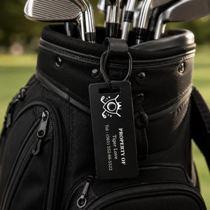 May include: Black golf bag with golf clubs inside. A black tag with white text and a golf-themed logo is attached to the bag. The tag says "PROPERTY OF Tiger Love" and a phone number.
