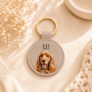 May include: A personalized keychain featuring a dog's portrait. The round, light gray leather pendant has a gold-colored ring and the name "RAY" printed on it. The dog has long, brown fur and is wearing a collar.