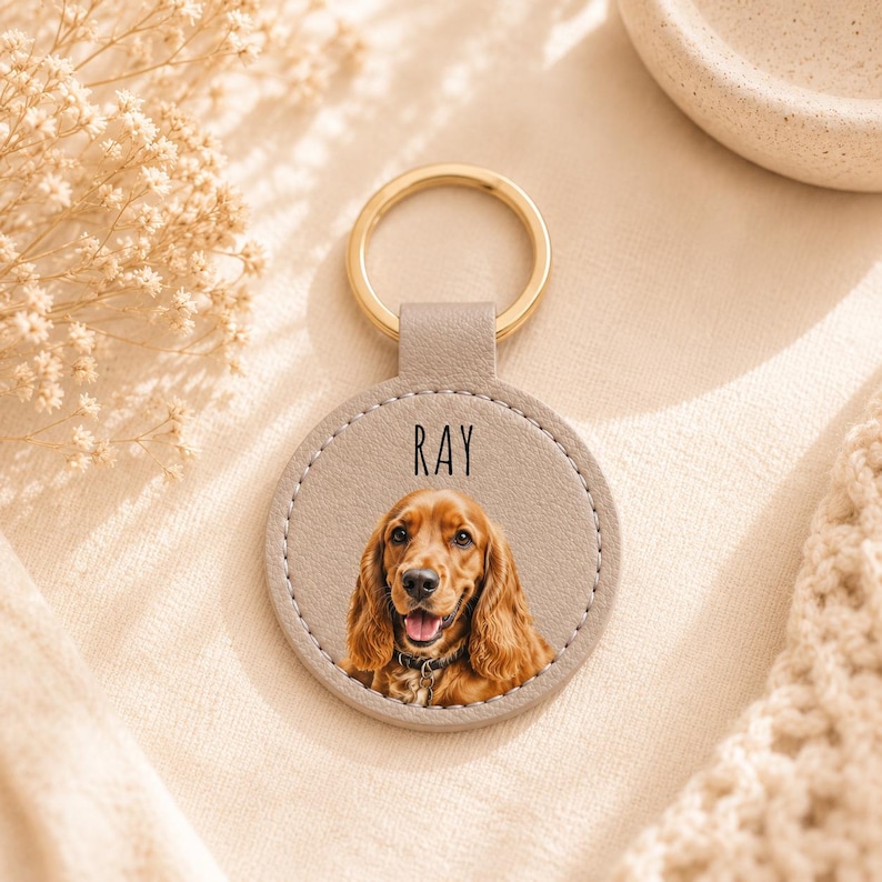 May include: A personalized keychain featuring a dog's portrait. The round, light gray leather pendant has a gold-colored ring and the name "RAY" printed on it. The dog has long, brown fur and is wearing a collar.