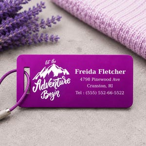May include: A purple luggage tag with white text reading "Let the Adventure Begin" and a mountain graphic. The tag displays a name, address, and phone number. A purple cable and lavender sprigs are also visible.