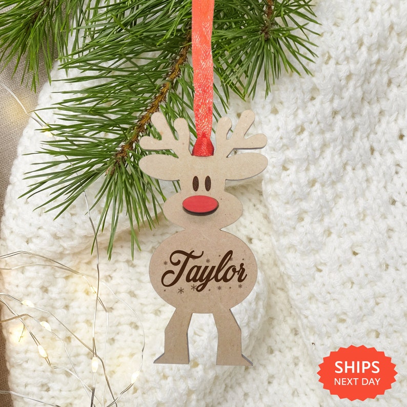 May include: A light brown wooden reindeer ornament featuring a red nose and the name Taylor engraved in dark brown script. The ornament is suspended by a bright red ribbon and includes small engraved snowflake details below the name.