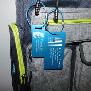 May include: Two blue luggage tags with a white American flag design. The tags have the name Emmett Brown, an address, an email address, and a phone number. The tags are attached to a gray and black backpack with a green zipper.