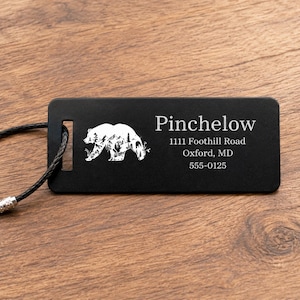 May include: Black luggage tag featuring a white bear silhouette. The tag displays the name "Pinchelow" and address details: 1111 Foothill Road, Oxford, MD 555-0125. A black cord is attached for securing the tag.