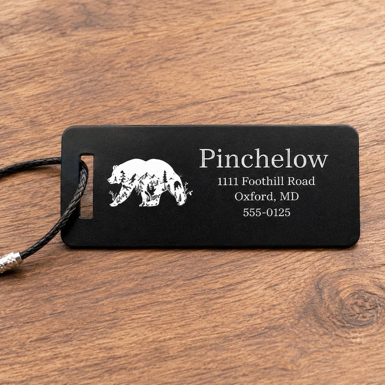 May include: Black luggage tag featuring a white bear silhouette. The tag displays the name "Pinchelow" and address details: 1111 Foothill Road, Oxford, MD 555-0125. A black cord is attached for securing the tag.