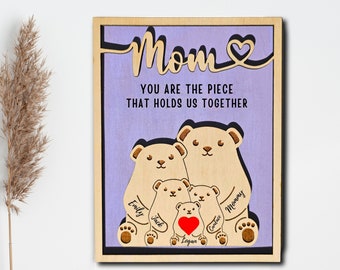 May include: Wooden sign with a purple background and the text "Mom You are the piece that holds us together". The sign features a family of bears with the names "Mommy", "Cadence", "Logan", "Jake", and "Emily" written on them.