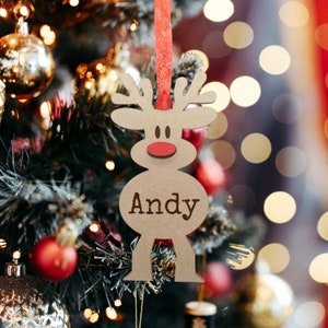 May include: A light brown wooden reindeer ornament featuring a red nose and the name Andy engraved in dark brown letters. The ornament is suspended by a thin red ribbon and has a cutout design shaped like a stylized reindeer with antlers.