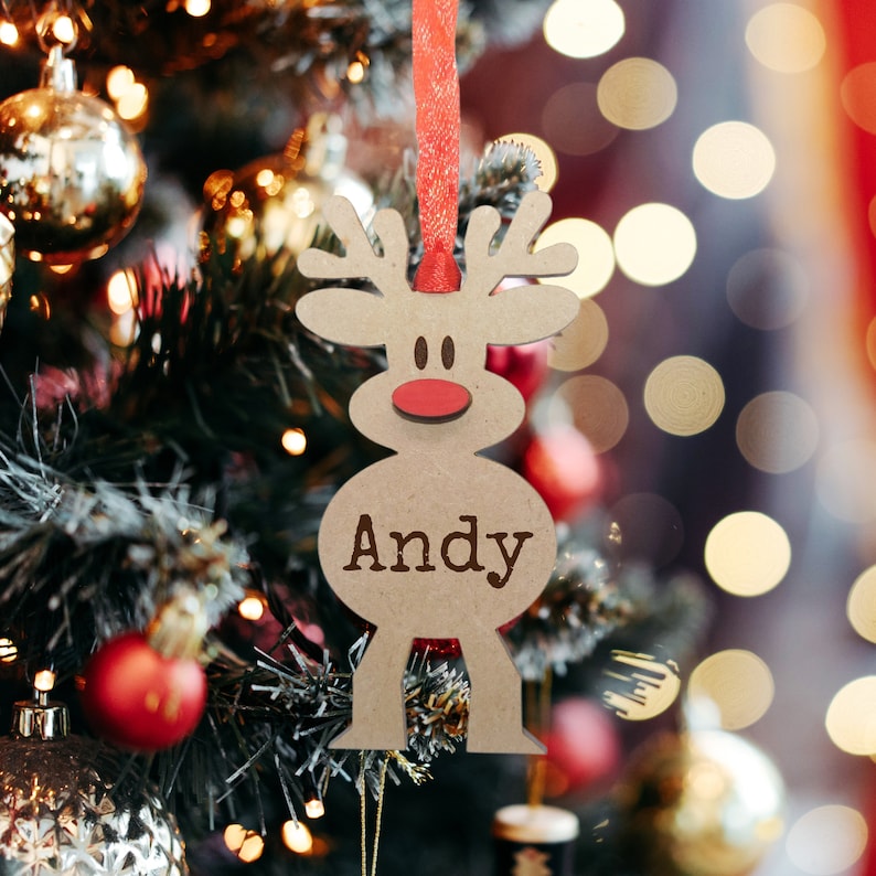 May include: A light brown wooden reindeer ornament featuring a red nose and the name Andy engraved in dark brown letters. The ornament is suspended by a thin red ribbon and has a cutout design shaped like a stylized reindeer with antlers.