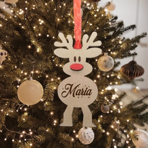 May include: A light-colored wooden reindeer ornament featuring a bright red nose and the name Maria printed in dark script lettering. The ornament is suspended by a textured red ribbon and displays a stylized antler design at the top.