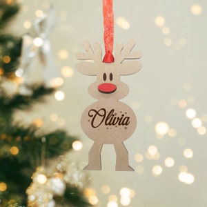 May include: A light brown wooden reindeer ornament featuring a red nose, dark brown eyes, and the name Olivia engraved in a script font on its belly. The ornament is suspended by a red ribbon and includes small snowflake accents.