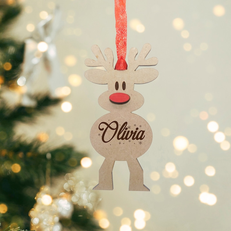 May include: A light brown wooden reindeer ornament featuring a red nose, dark brown eyes, and the name Olivia engraved in a script font on its belly. The ornament is suspended by a red ribbon and includes small snowflake accents.