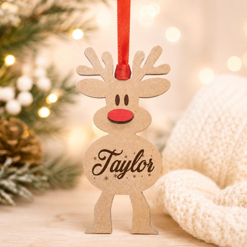 May include: A light brown wooden reindeer ornament featuring a bright red nose and the name Taylor engraved in dark brown script on its belly. The ornament is suspended by a thin red ribbon and includes small engraved snowflake accents.