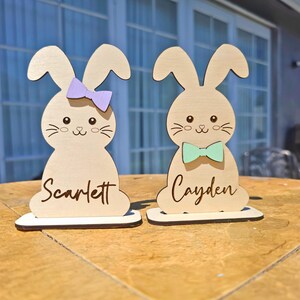 Op de afbeelding: Twee houten paashaasdecoraties. E&eacute;n konijn heeft een paarse strik en de naam "Scarlett" erop geschreven. Het andere konijn heeft een mintgroene strik en de naam "Cayden" erop geschreven. Beide konijnen hebben een schattig gezicht.
