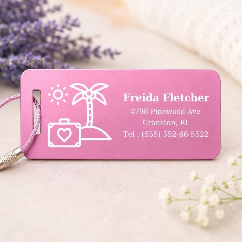 May include: A pink luggage tag featuring a white graphic of a sun, palm tree, and suitcase with a heart. The tag displays the name "Freida Fletcher" and contact details. A silver cable and lavender sprigs are visible.