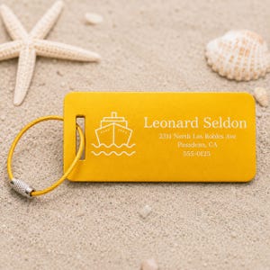 May include: A yellow luggage tag featuring a cruise ship icon and the name "Leonard Seldon" in white. The tag displays an address and phone number. A gold wire loop is attached. The background is sand with a starfish and seashell.