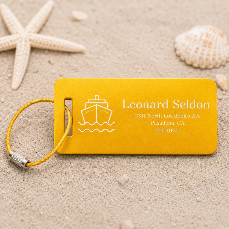 May include: A yellow luggage tag featuring a cruise ship icon and the name "Leonard Seldon" in white. The tag displays an address and phone number. A gold wire loop is attached. The background is sand with a starfish and seashell.