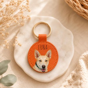 May include: Orange leather keychain with a gold ring. The keychain features a photo of a dog with the name "LUNA" printed in black. The dog has tan fur, a black nose, and blue collar.