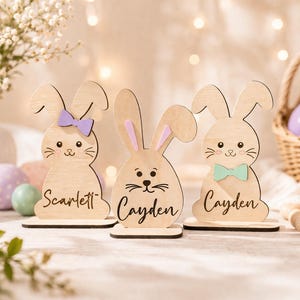Op de afbeelding: Drie houten paashaasdecoraties met namen, elk met een verschillend gekleurde vlinderdas of strik. De konijnen zijn lichtbruin met zwarte gezichtskenmerken en staan op kleine houten voetstukken. De namen "Scarlett" en "Cayden" staan op de konijnen.