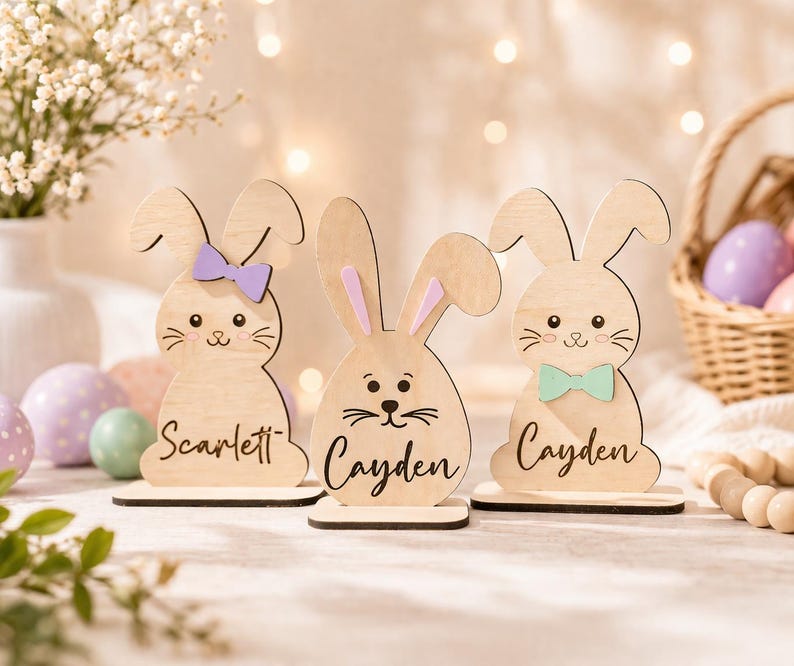 Op de afbeelding: Drie houten paashaasdecoraties met namen, elk met een verschillend gekleurde vlinderdas of strik. De konijnen zijn lichtbruin met zwarte gezichtskenmerken en staan op kleine houten voetstukken. De namen "Scarlett" en "Cayden" staan op de konijnen.