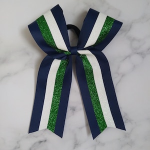 May include: A hair bow constructed from grosgrain ribbon featuring a layered design with navy blue, white, and green glitter stripes. The bow is secured with a central knot and attached to a black elastic hair tie, forming a symmetrical shape.