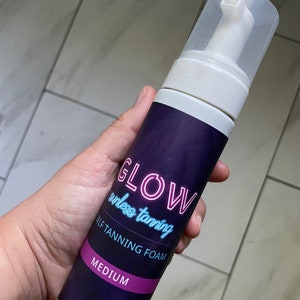 May include: A white bottle of self-tanning foam with a purple label that says "GLOW sunless tanning SELF TANNING FOAM MEDIUM 7 FL OZ (207 ml)".