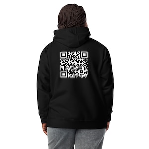 May include: Black hoodie featuring a white QR code on the back. The hoodie has a hood and a drawstring. The person is wearing grey trousers. The QR code is a square pattern of black and white squares.