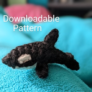May include: A black and white crocheted orca whale plush toy on a blue fuzzy surface. The text "Downloadable Pattern" is visible above the toy.