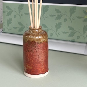 Puede incluir: Un difusor de caña de cerámica con un cuerpo marrón rojizo texturizado y una parte superior marrón. La base es blanca. Varias cañas de color claro se insertan en la abertura. El fondo presenta un patrón de hojas verdes y blancas y el texto "littleacornpottery.co.uk | @ f Etsy".