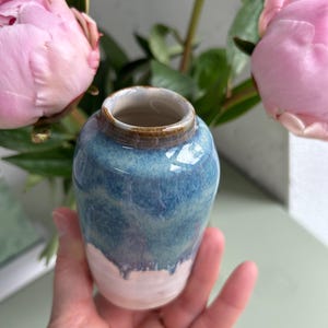 Może przedstawiać: Mały, ręcznie robiony ceramiczny wazon z niebiesko-białą glazurą. Wazon ma zaokrąglony kształt i wąski otwór. Niebieska glazura przechodzi od ciemniejszego odcienia u góry do jaśniejszego odcienia u dołu. Różowe piwonie w tle.