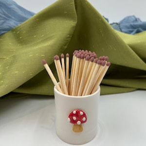 May include: A white ceramic cylindrical holder containing a bundle of wooden matches with brown tips. The holder features a small, raised, three-dimensional red mushroom decoration with white spots on its front surface.