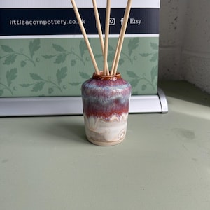 Puede incluir: Un pequeño jarrón difusor de caña de cerámica con un degradado de colores, incluyendo blanco, rojo y azul. El jarrón contiene varias cañas de madera de color claro. El fondo presenta el texto "LITTLE ACORN POTTERY" y la dirección del sitio web.